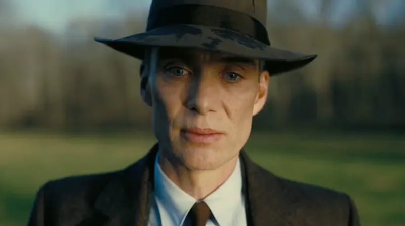 A person wearing a dark suit, tie, and wide-brimmed hat stands outdoors in front of blurred trees, looking ahead with a serious expression.