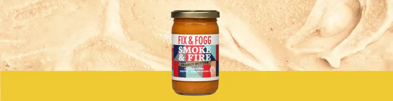 A jar of Fix & Fogg Smoke & Fire peanut butter stands against a background of creamy peanut butter with a yellow bottom border.