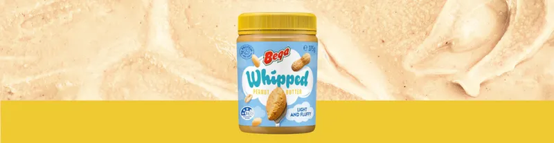 A jar of Bevi Whipped Peanut Butter sits against a creamy peanut butter background with a yellow stripe at the bottom. The jar features peanuts and blue and white text.