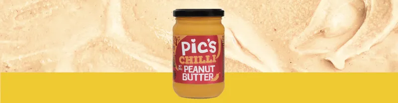 A jar of Pic’s Chilli Peanut Butter is centered on a yellow background with a close-up of peanut butter spread behind it.