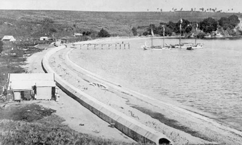 A black and white photo of a curved shoreline with a dock, a few boats, small buildings, and trees in the background. A long, narrow structure runs along the beach.