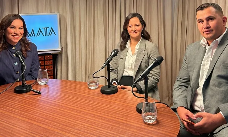 Three people in suits sit around a wooden table with microphones and glasses of water, ready for a discussion. A screen with the word “MATA” is visible in the background. All appear to be engaged and smiling.