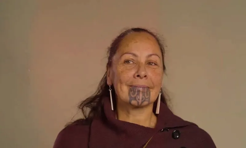 A woman with long dark hair, wearing a maroon shawl and long earrings, has a traditional Māori chin tattoo and is looking slightly to the side against a plain background.