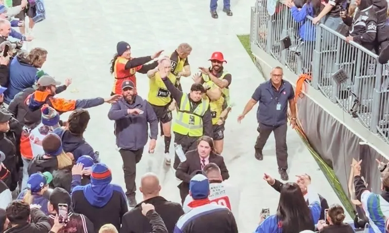 A crowd surrounds security escorting a man off a sports field. Stewards in yellow vests and staff in dark uniforms guide the man as fans watch and take photos from the stands.