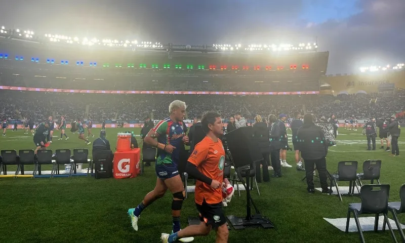 Rugby league players warm up on a stadium field under bright lights, with coaches and officials nearby. The stands are filled with spectators and the sky is cloudy.