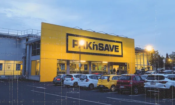 A yellow PAK’nSAVE supermarket building with several parked cars in front, taken during early evening with streetlights on.