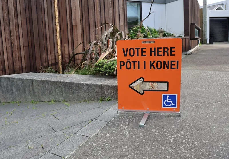 An orange sign reads “Vote Here, Pōti i konei” with an arrow pointing left. It includes a wheelchair accessibility symbol. The sign is on a sidewalk near a building.