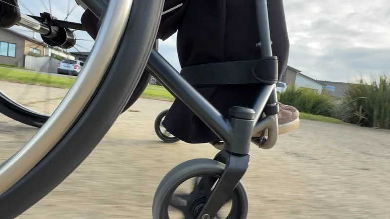 Close-up of a person’s foot on a wheelchair footrest, moving outdoors on a paved path, with houses, grass, and a cloudy sky in the background.