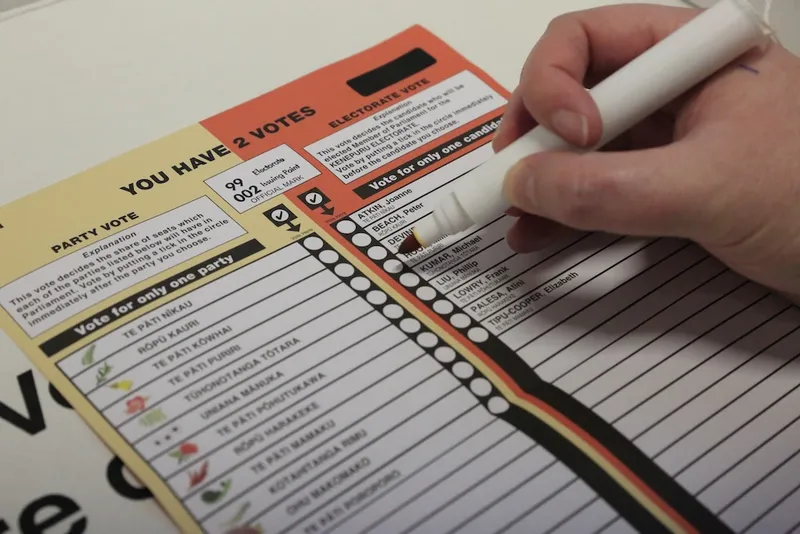 A hand is marking a ballot paper with a pen. The ballot lists political parties and candidates, with instructions indicating voters can select two options. The paper is colorful, including red, yellow, and black sections.
