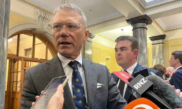 A man in a suit speaks to reporters holding microphones and phones in a formal building with columns and ornate ceilings. Another man stands behind him.