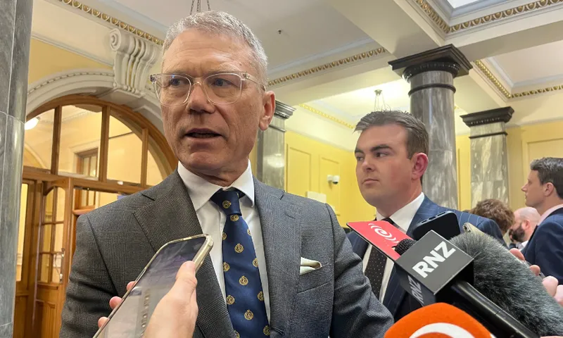 A man in a suit speaks to reporters holding microphones and phones in a formal building with columns and ornate ceilings. Another man stands behind him.