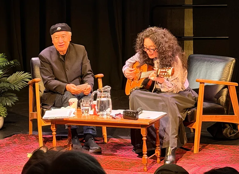an older man and younger woman sit in chairs on a stage. the man wears a kangol hat and the woman has curly hair and is playing a guitar