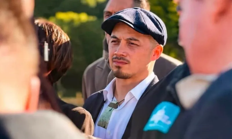 A man wearing a flat cap and a green pendant stands among a group of people outdoors, looking ahead with a serious expression. Trees and sunlight are visible in the blurred background.