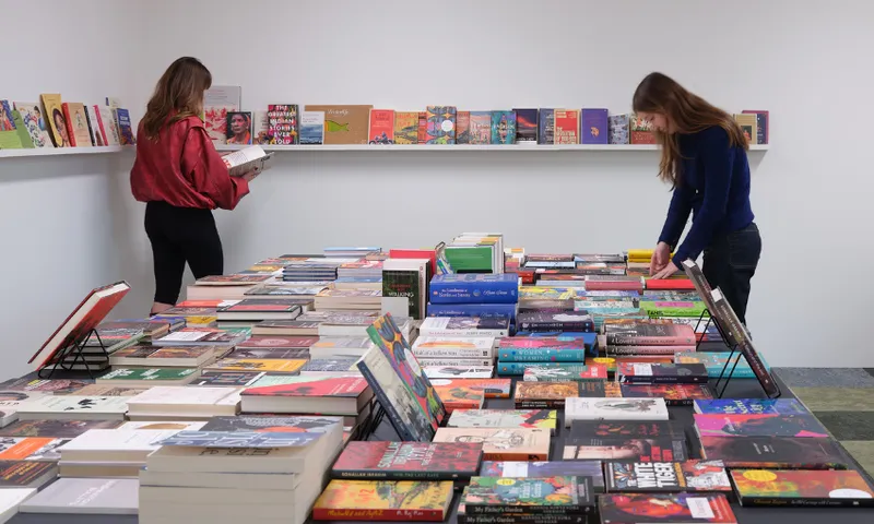 A table full of books is being browsed by two young women. There are shelves around the walls also covered in books.
