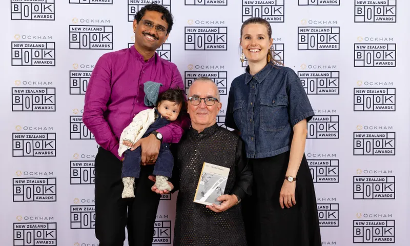 A man is holding a baby and standing next to another man who is holding a book. Beside them is a young woman. Behind them all is a branded wall showing the logo for the Ockham New Zealand Book Awards.