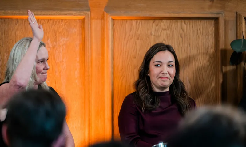 Two women sit side by side in front of a wooden panel; the woman on the left raises her hand, while the woman on the right, wearing a maroon top, looks to her left with a slight smile.