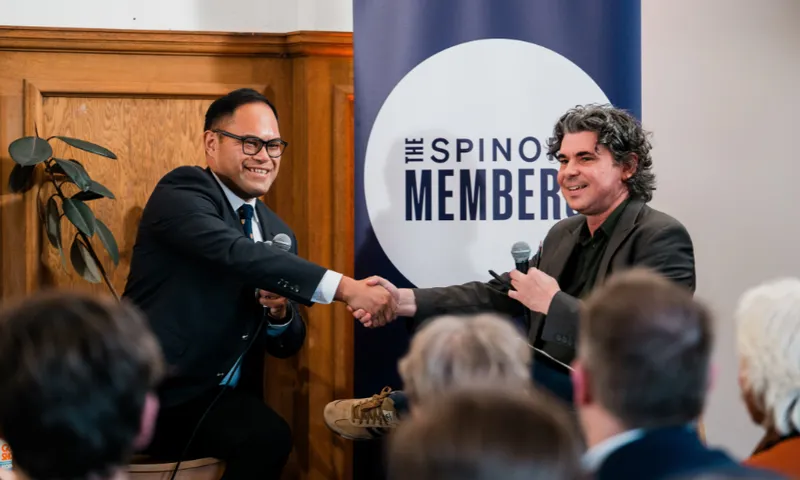Two men sit and shake hands onstage during a discussion event, holding microphones. An audience watches in the foreground. A banner behind them reads “The Spinoff Members.”.