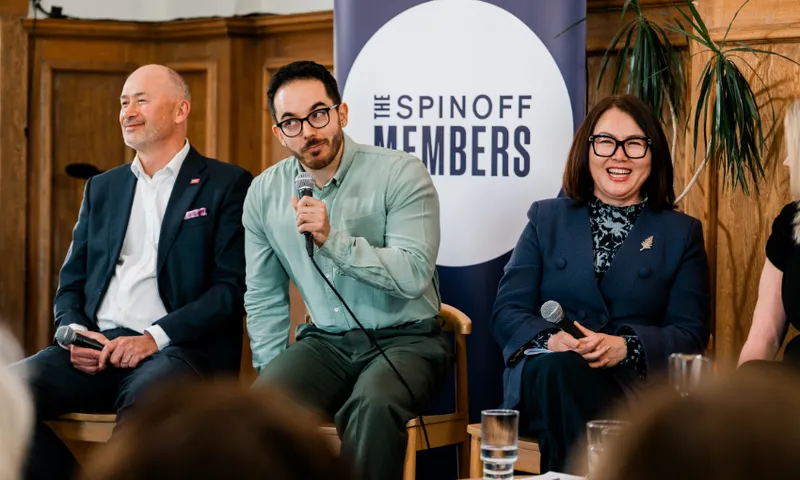 Three people sit on chairs holding microphones at a panel event, smiling and engaged, with “The Spinoff Members” sign in the background and wooden paneling behind them.