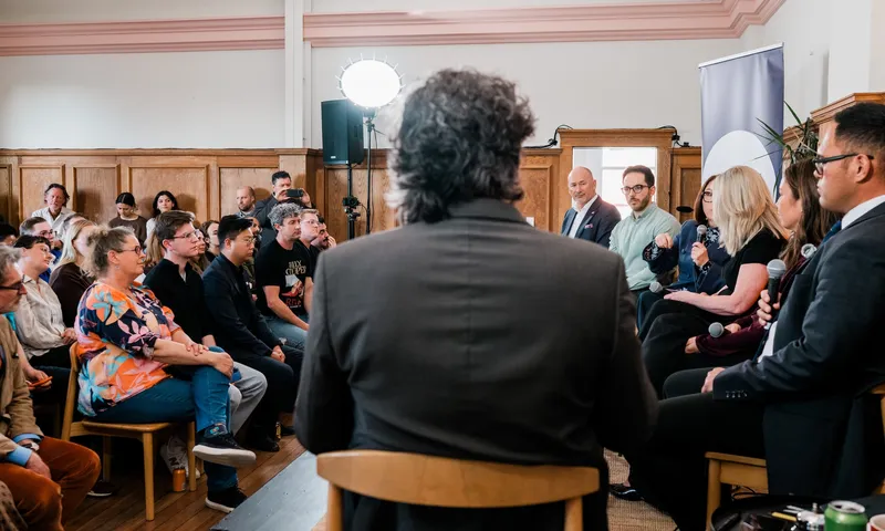 A group of panelists speaks to an audience in a wood-paneled room. People are seated, listening attentively. A man with curly hair is in the foreground, seen from behind. Bright lights and a banner are in the background.