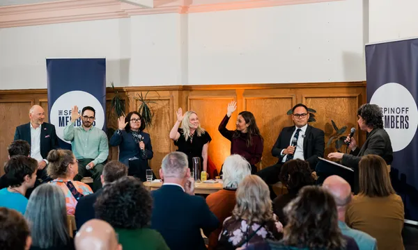 six adults in business casual attire sit on a stage as a panel, with a suited man as the moderator. three women and one man have their hands raised while two men don't