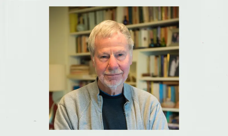 an older man in a collared shirt stares down the lens with a large bookshelf behind him