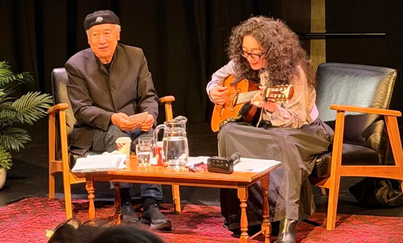 an older man and younger woman sit in chairs on a stage. the man wears a kangol hat and the woman has curly hair and is playing a guitar