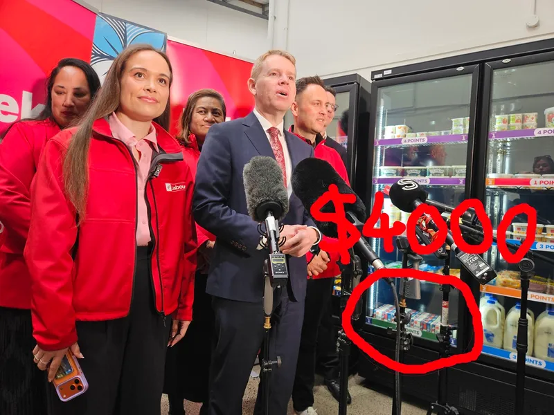 A group of people in red jackets stands by a man speaking into microphones at a supermarket, with a fridge behind them and “$4000” written in red over dairy products in the background.