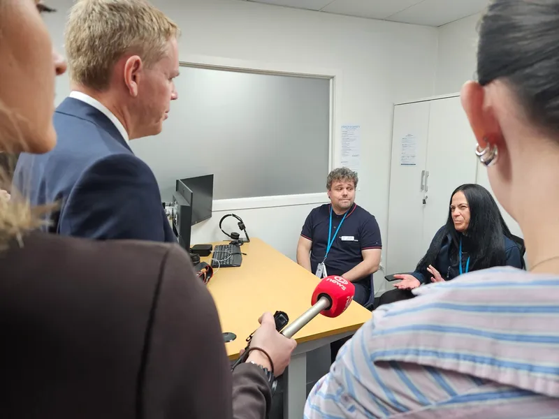 Four people talk in an office. Two sit at a table, one with a lanyard gestures while speaking. Two others, one holding a red microphone, stand with their backs to the camera. Computer monitors and papers are on the table.
