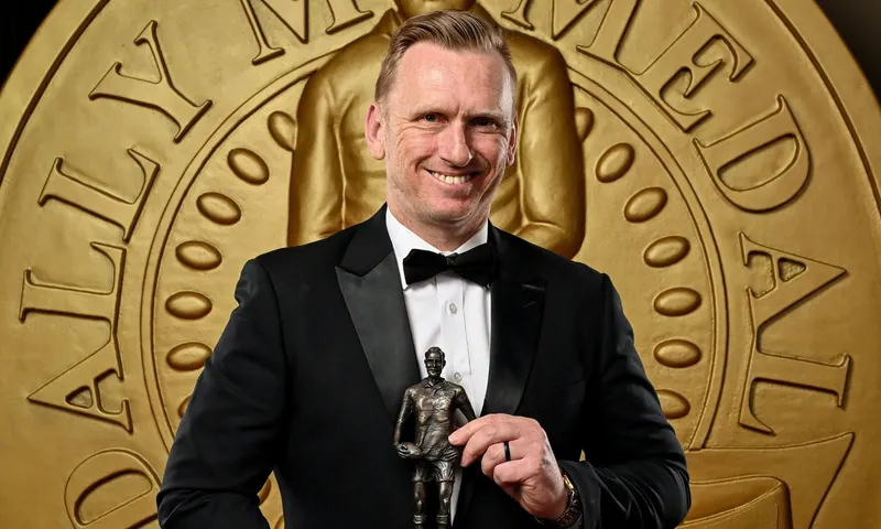 A man in a black tuxedo smiles while holding a small statue award in front of a large gold medal backdrop.