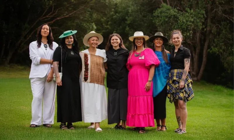 Seven women stand together outdoors on green grass, smiling. They wear a mix of colourful and neutral outfits, including dresses, hats, and accessories, with trees and bushes in the background.