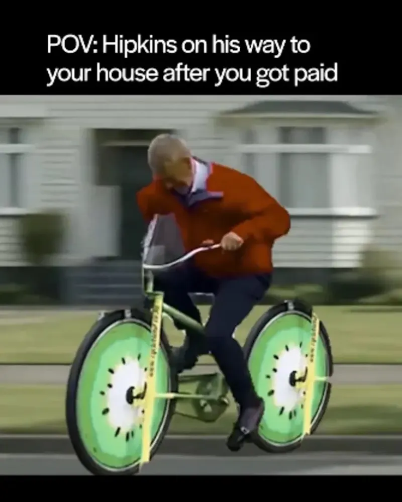 A man in a red jacket rides a bike with wheels resembling sliced kiwis. Text above reads, “POV: Hipkins on his way to your house after you got paid.”.