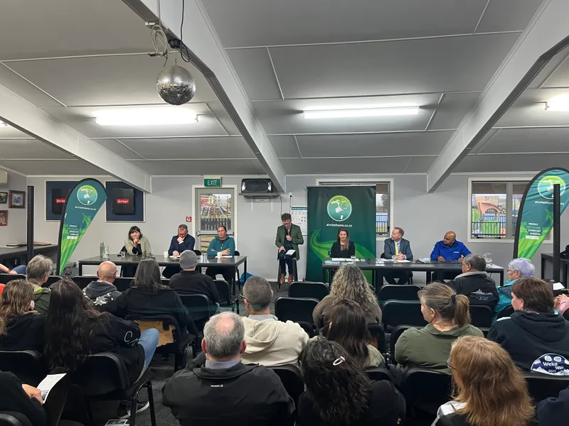 a brightly lit community room with two tables where six political candidates sit, with a moderator on a stool in the middle. a small audience watches them