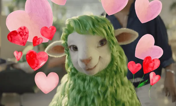 A smiling cartoon sheep with green wool is surrounded by red and pink heart illustrations. The background is a softly blurred indoor setting.
