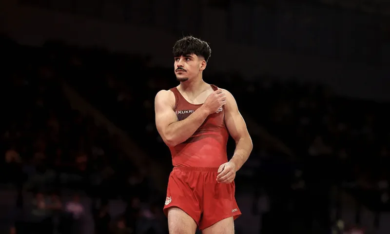 a male gymnast in a red competition uniform stands looking off to the side with a dark, blurred background behind
