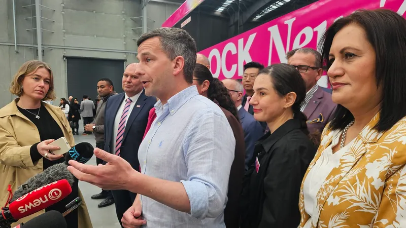 David Seymour, a middle aged NZ politician with greying hair stands in front of a sceptical-looking reporter and a bright pink bus at the Act campaign launch in a large building.