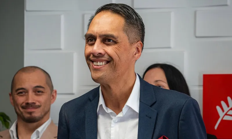 A smiling man in a navy suit and white shirt stands at an indoor political event, with two people behind him and a red Labour Party sign visible to the right.
