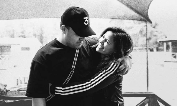 A young man in a black cap has his arm around Huia O’Sullivan as she smiles up at him in a black-and-white photograph.