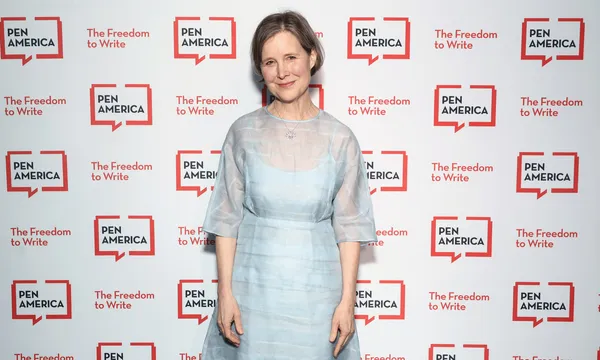 A woman in a light blue dress standing against a background with the repeated logo of PEN America.