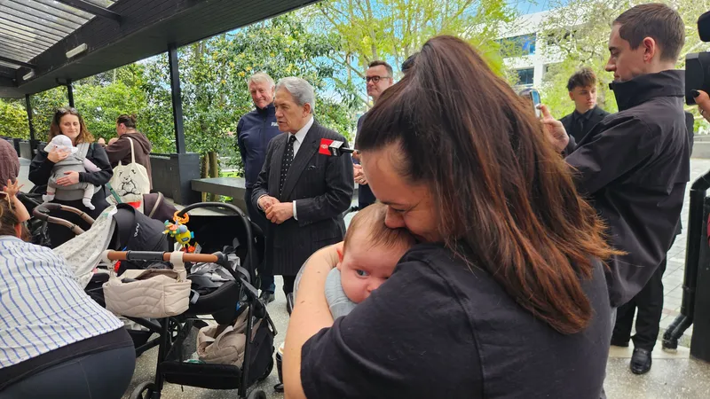 A woman holds a baby whose face is turned away as NZ politicians Winston Peters stands in the background with grey hair and a dark suit.