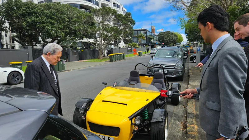 Winston Peters, a grey-haired NZ politician in a dark suit, looks at a bright yellow fibreglass car. 