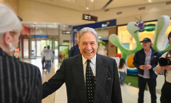 Winston Peters, a smiling man in a suit and tie, stands facing the camera with his arm held out, in a mall setting. A woman with white hair is a the left of the image, back on, and a photographer can be seen at the right.