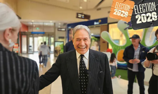 Winston Peters, a smiling man in a suit and tie, stands facing the camera with his arm held out, in a mall setting. A woman with white hair is a the left of the image, back on, and a photographer can be seen at the right.