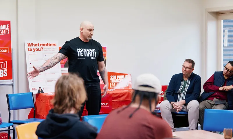 Mananui Ramsden wearing a “Honour Te Tiriti” T-shirt speaks to a group at an indoor campaign event.