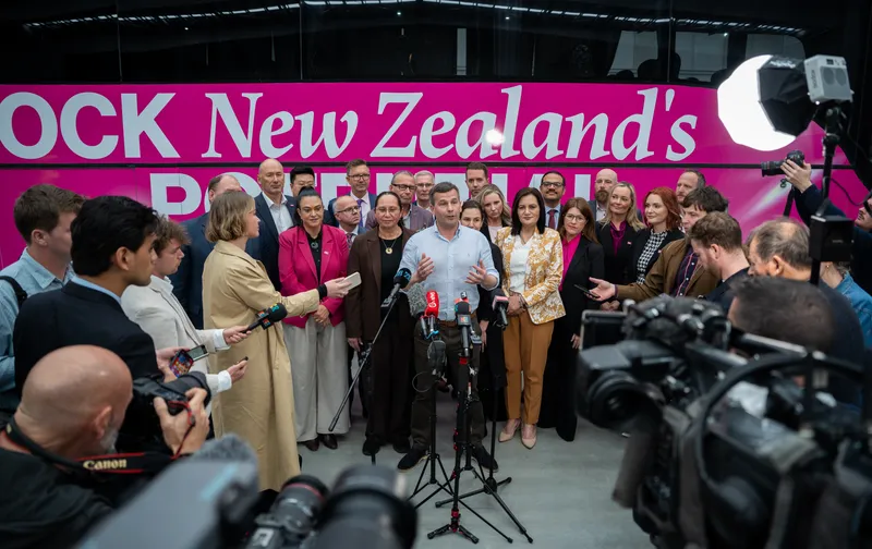 A media scrum surrounds a man, David Seymour, fronting a crowd of his peers and speaking to a microphone. They stand in front of a pink campaign bus promoting the Act Party.