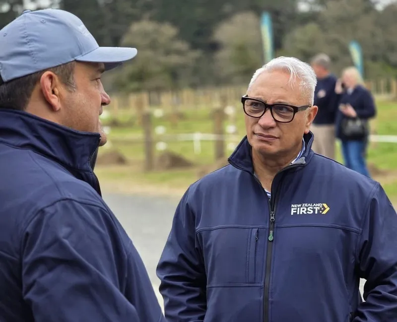 Taine Randell and Alfred Ngaro, wearing dark blue jackets with the NZ First logo on, stand in conversation in an outdoor setting. One wears a cap, the other wears dark-rimmed glasses.