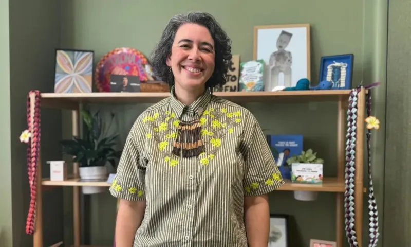 A middle-aged woman stands smiling in her office. There are photo frames and plants on the shelves behind her.