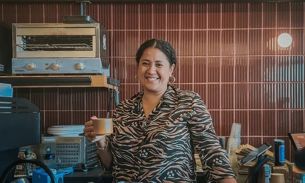 a woman with brown skin and brown tied-up hair stands in a cafe setting with brown tiles on the walls behind her. She's smiling and holding a coffee mug