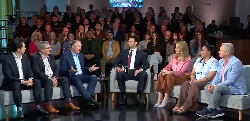 a panel of debaters sit on couches with the moderator in the middle and a small crowd behind them. A man in a navy suit with a blue shirt speaks enthusiastically