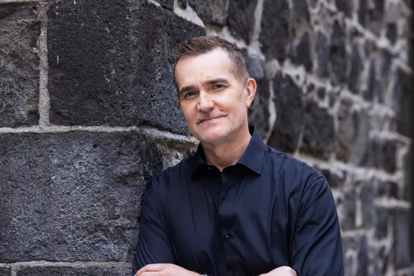John Aiken wears a black polo shirt and leans against a stone building, staring into the camera with his ams folded