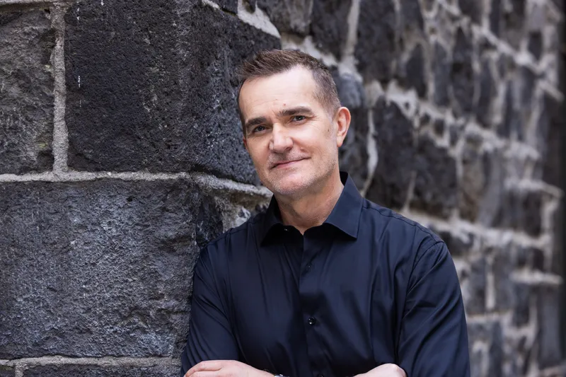 John Aiken wears a black polo shirt and leans against a stone building, staring into the camera with his ams folded